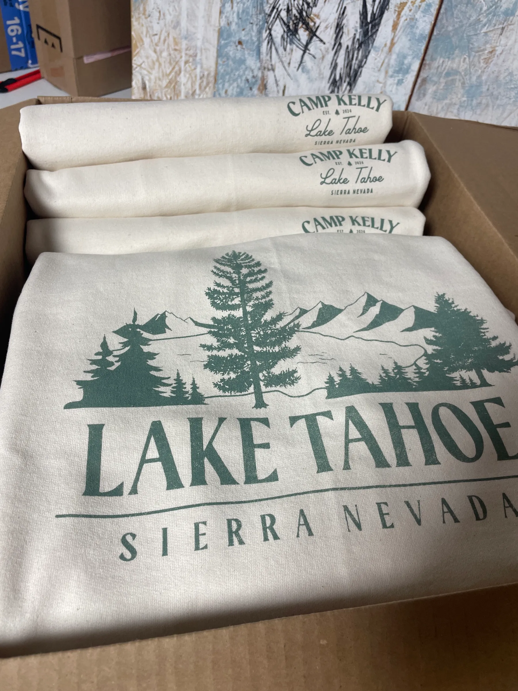 Natural-colored sweatshirts folded in a shipping box, printed with a green Lake Tahoe mountain and pine scene