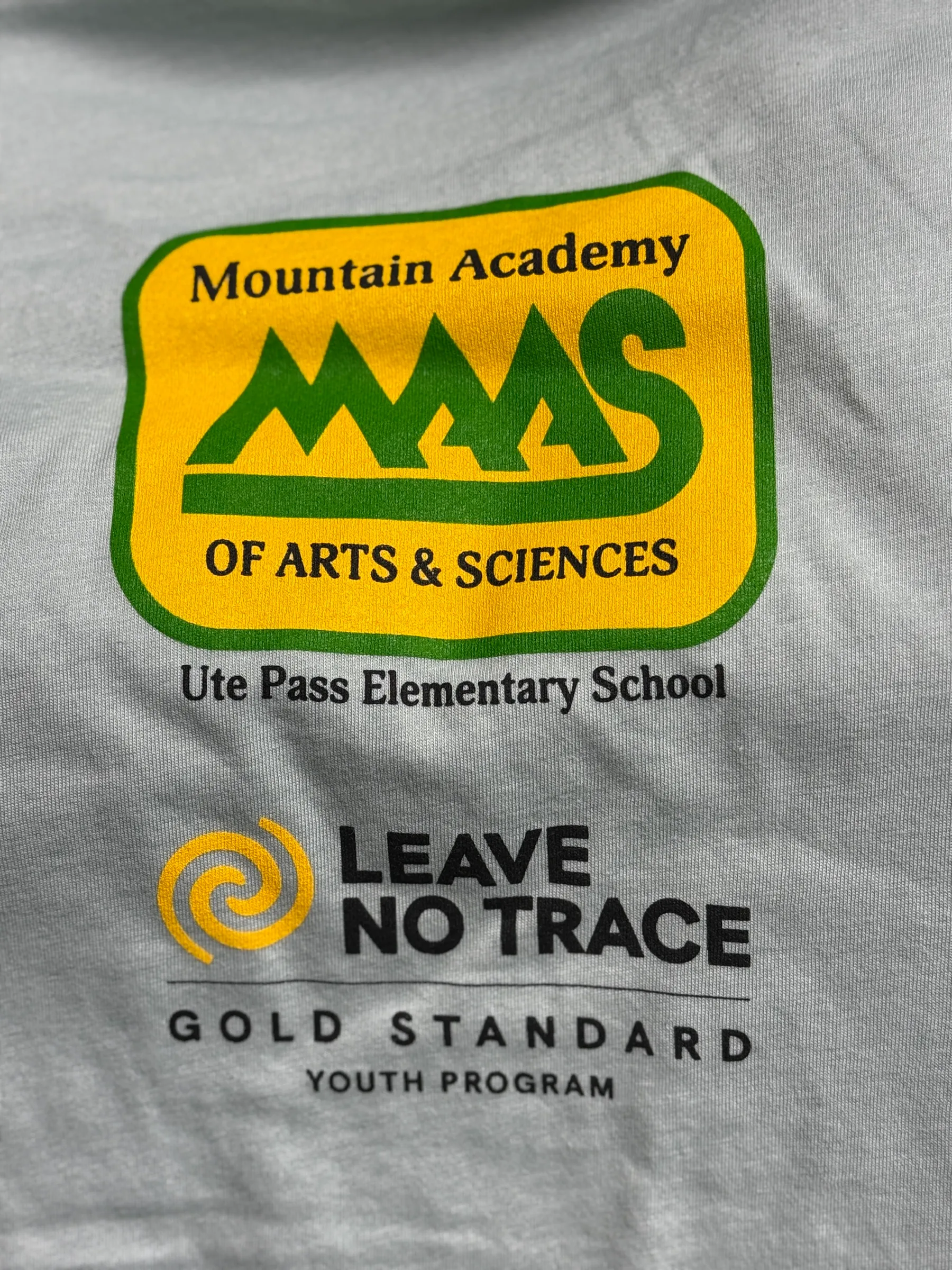 A pale green tee printed with the Mountain Academy of Arts & Sciences badge above a Leave No Trace Gold Standard Youth Program lockup