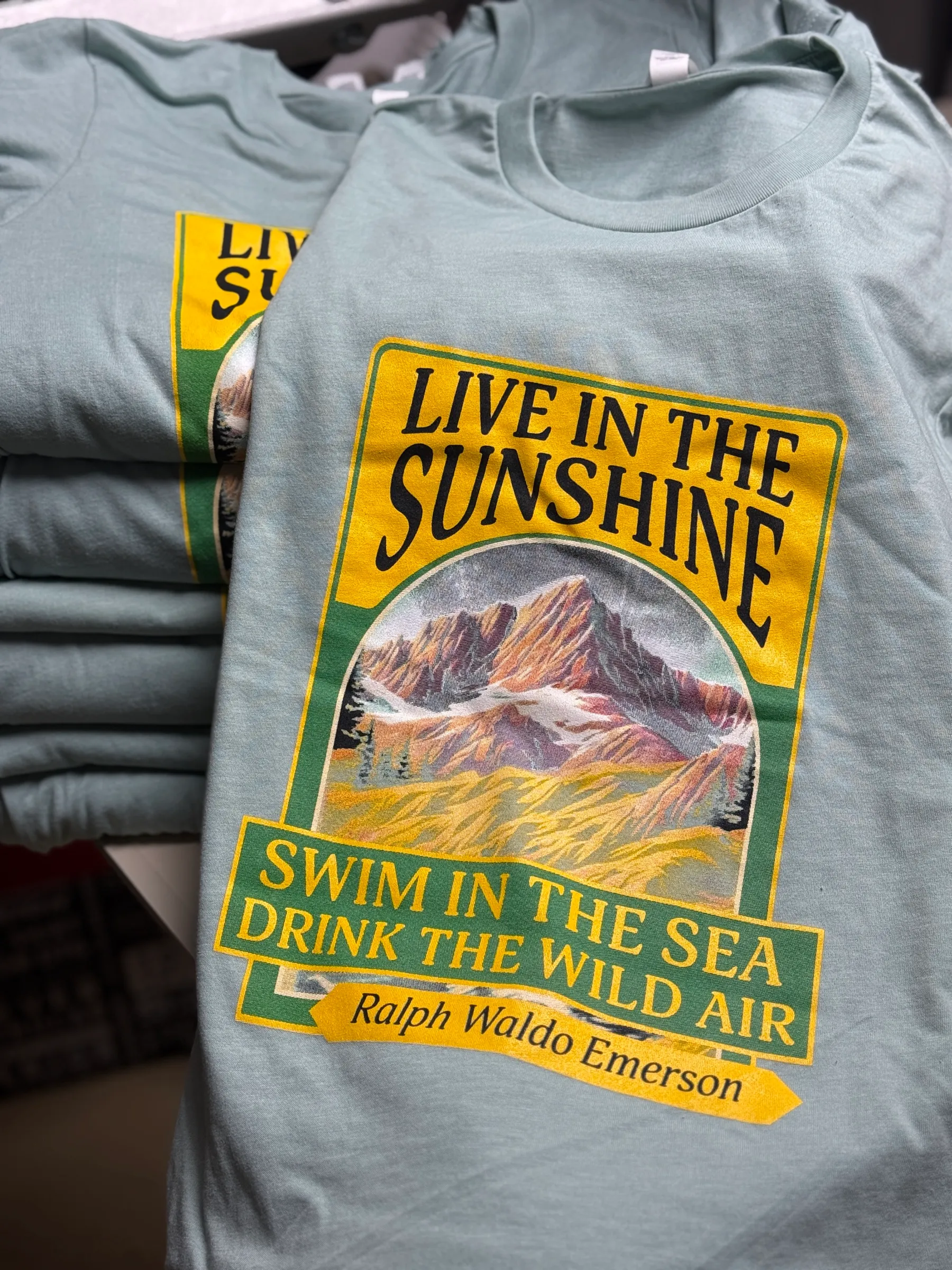 Sage green tees printed with a vintage-style Live in the Sunshine badge in yellow and green
