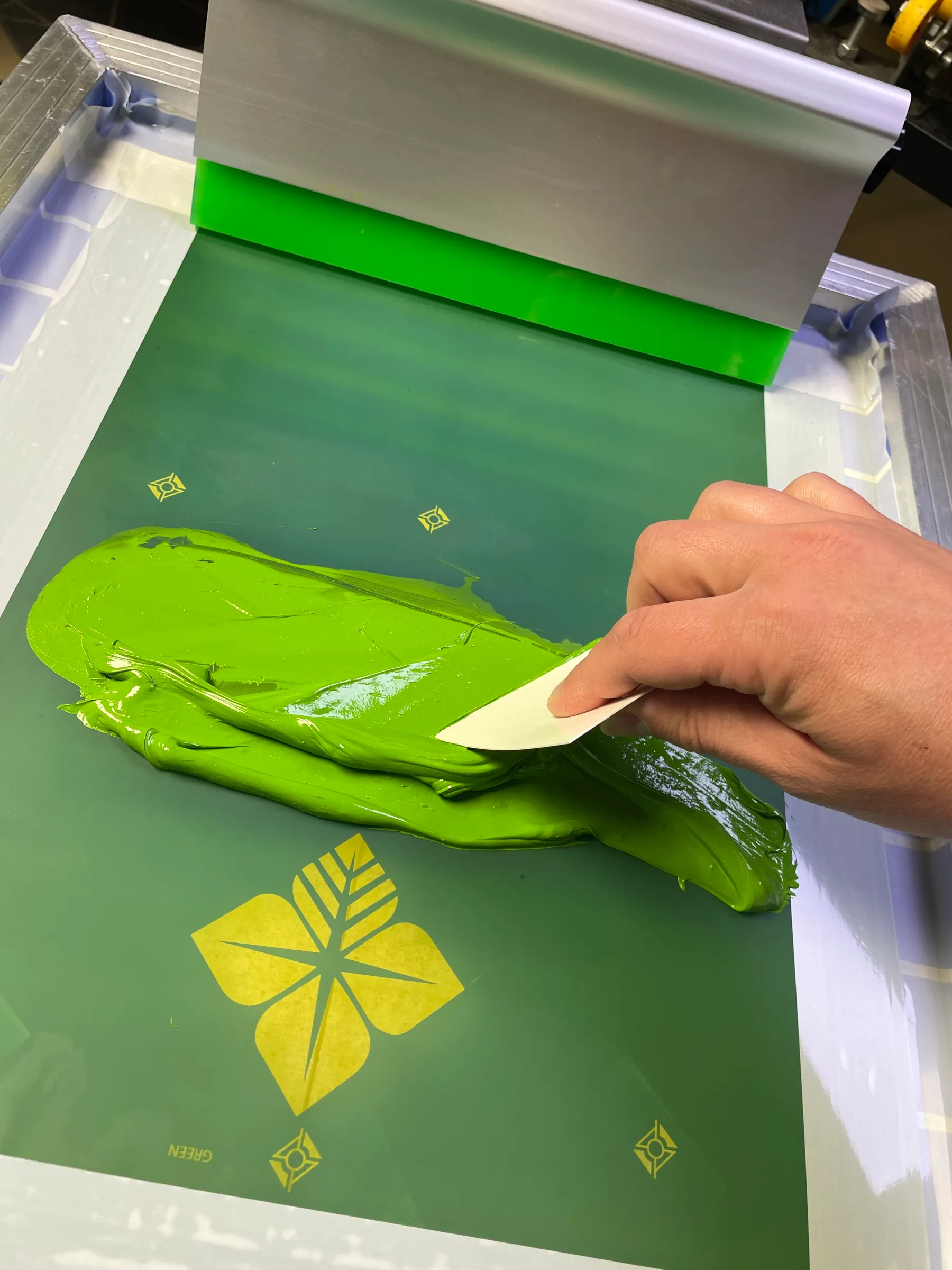 A hand pulling a card of bright green ink across a screen on the press, with the registration marks and the Farm Credit four-leaf mark visible in the mesh
