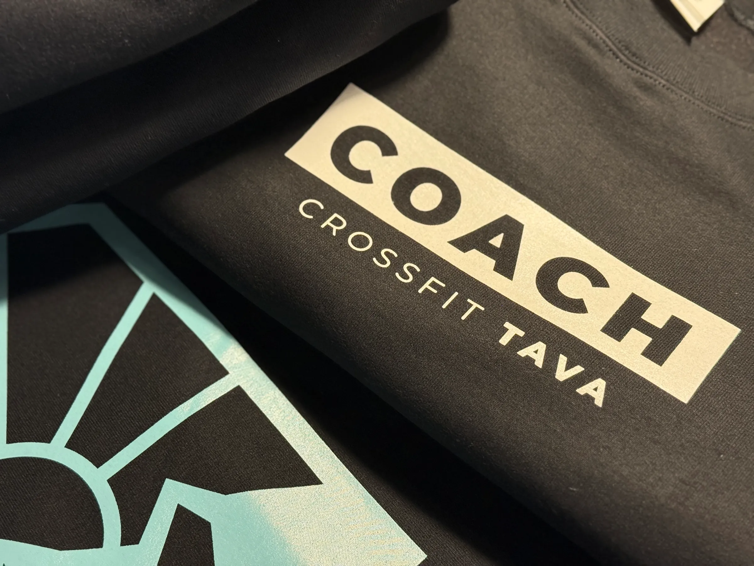 Close-up of a black garment printed with a solid block reading COACH above CROSSFIT TAVA in a lighter ink