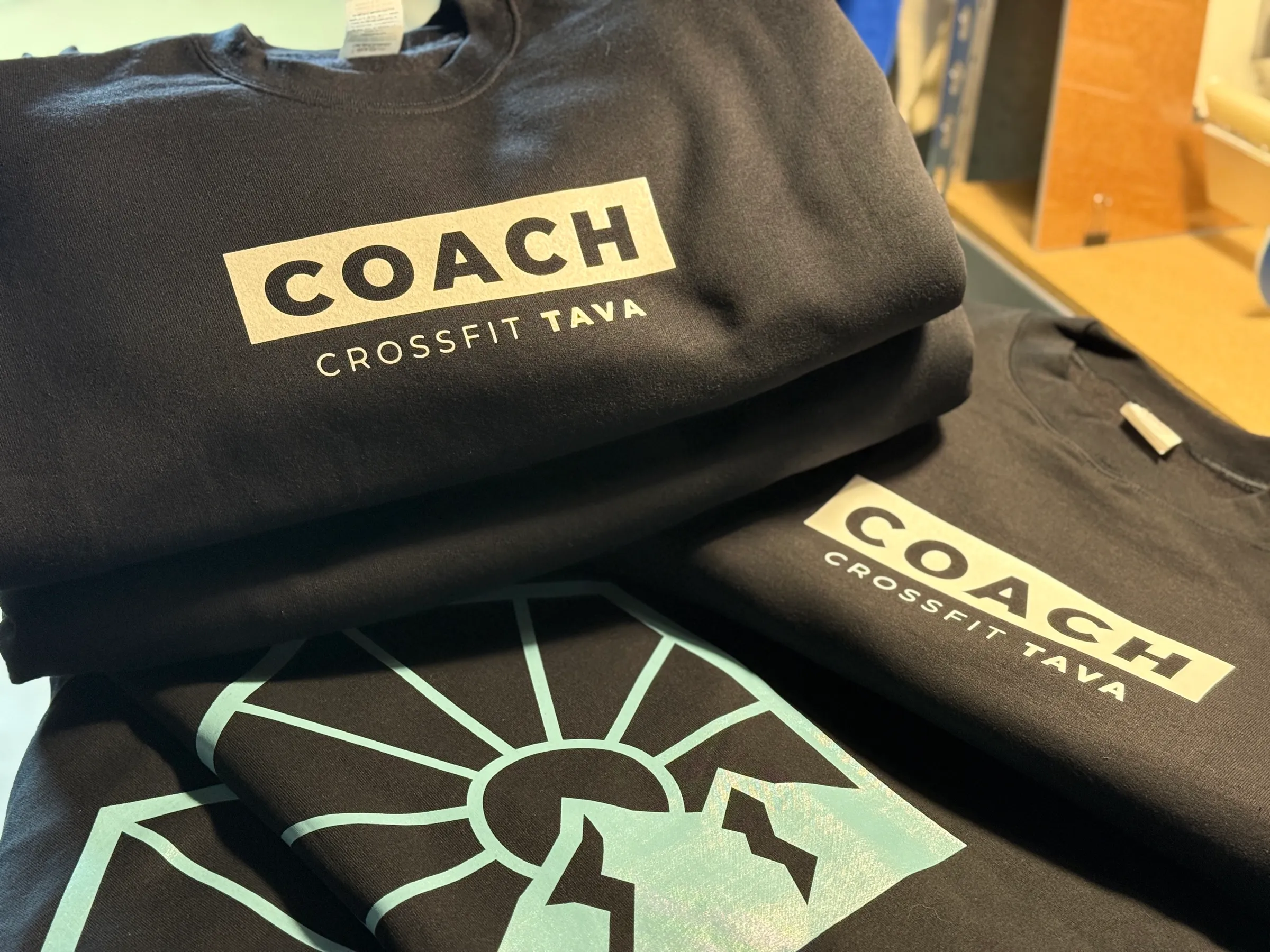 A stack of black CrossFit TAVA sweatshirts and tees printed with a pale mint sun-and-mountain mark