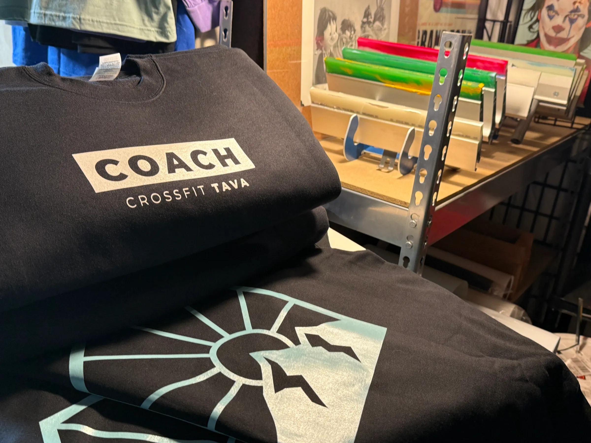 Folded black CrossFit TAVA garments on a work table beside racked screens and squeegees
